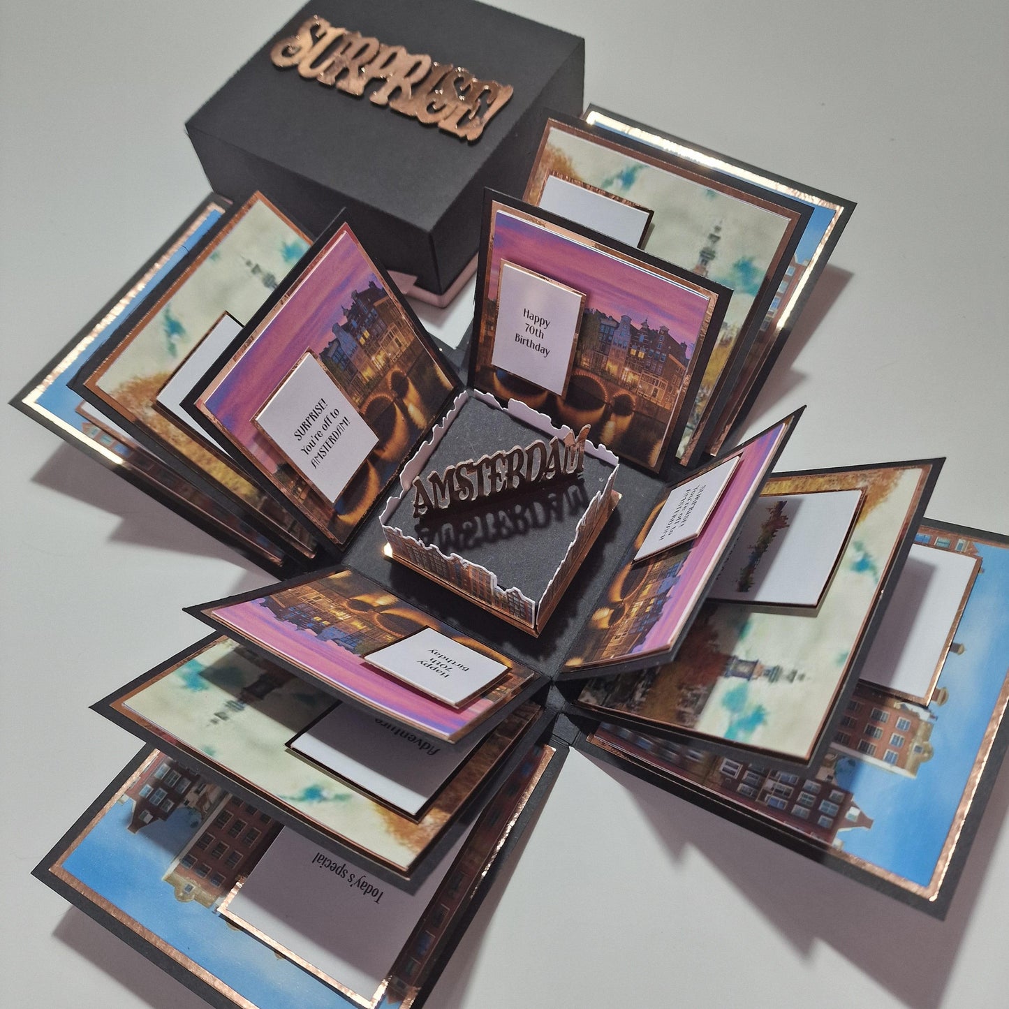 Exploding Occasions Box - Trip Reveal AMSTERDAM, featuring a black box with gold text, Amsterdam-themed paper panels, and cards detailing a surprise trip, ideal for special celebrations.