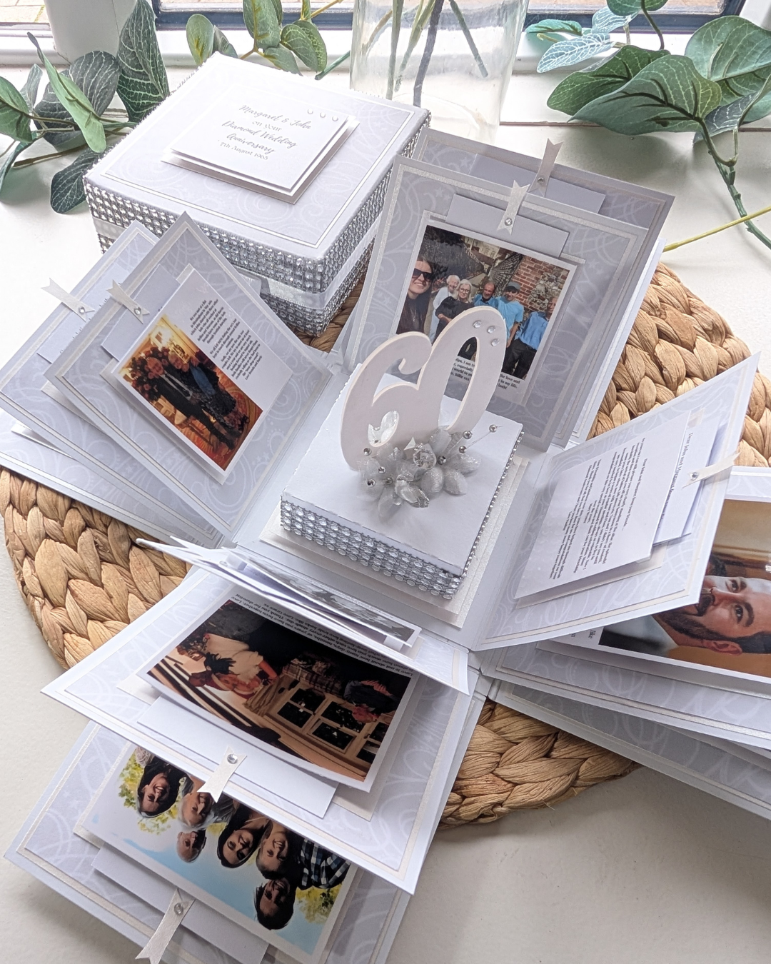 Open photo album, exploding box with family photos and a decorative heart on a woven mat.
