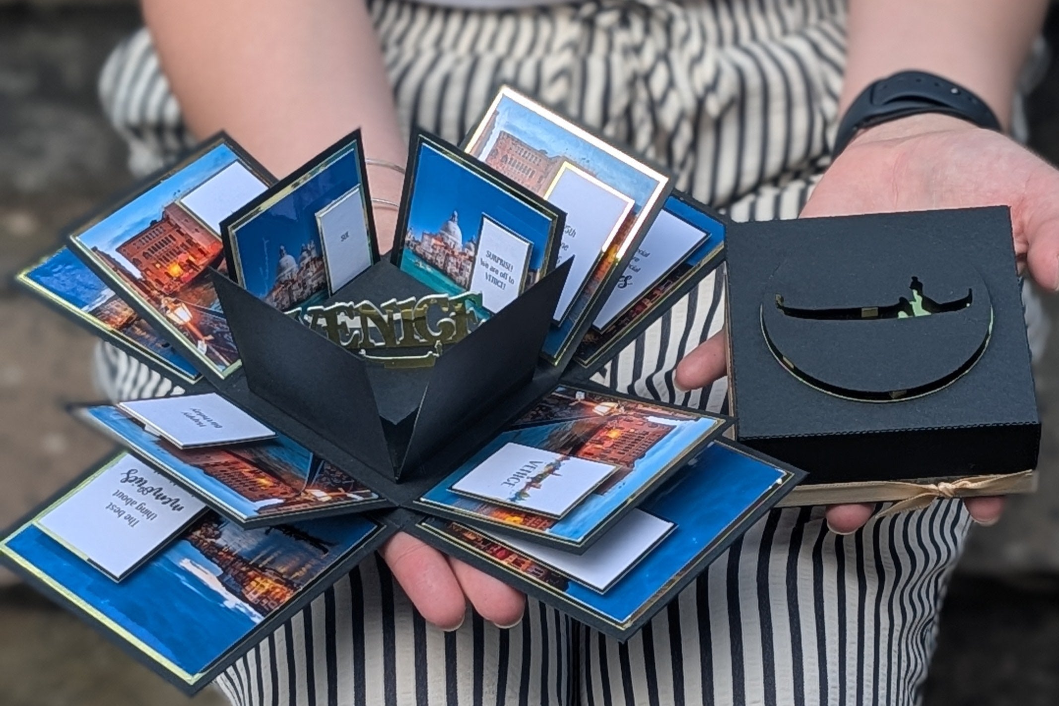 Person holding a black box with multiple cards inside, a trip reveal for a Venice vacation product, on a blurred background.