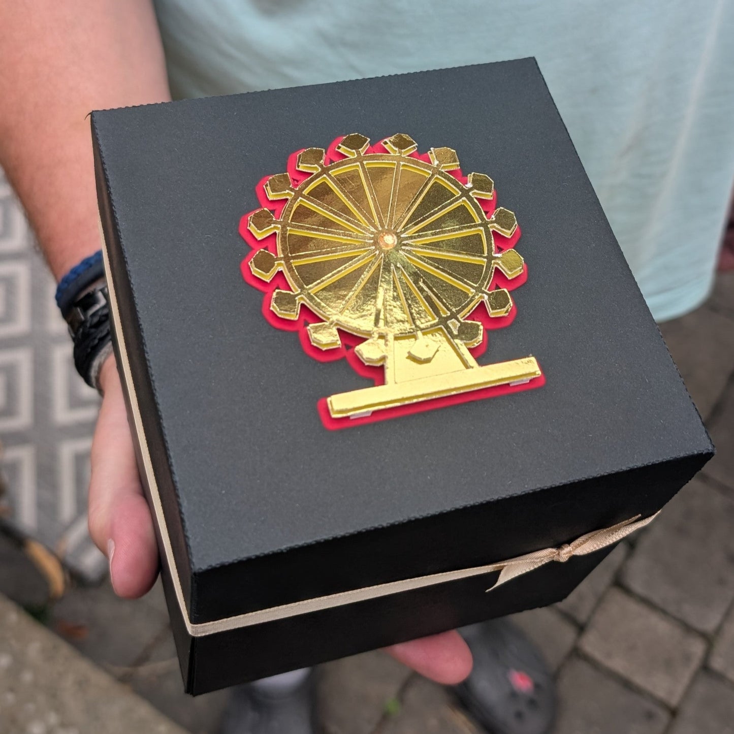 London - Special Occasion | Celebration Trip Reveal Explosion Box: A small black gift box with a gold-and-red Ferris-wheel design on the lid and a beige ribbon, ideal for announcing trips.