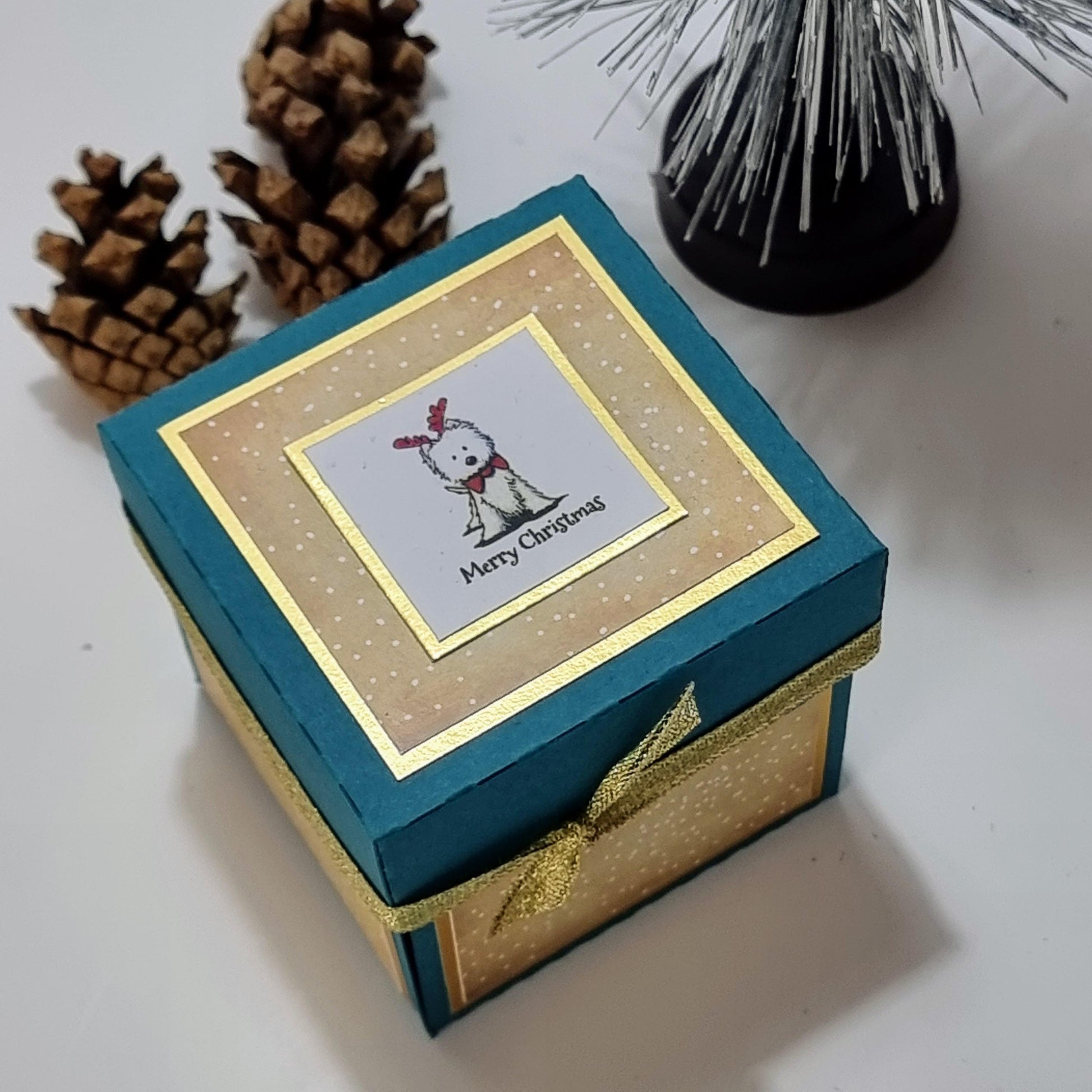 Lou Lou The Westie Christmas Candle Box features a teal box with gold accents, adorned with a West Highland Terrier illustration, pine cones, and a ribbon, containing a candleholder with a festive scented candle.
