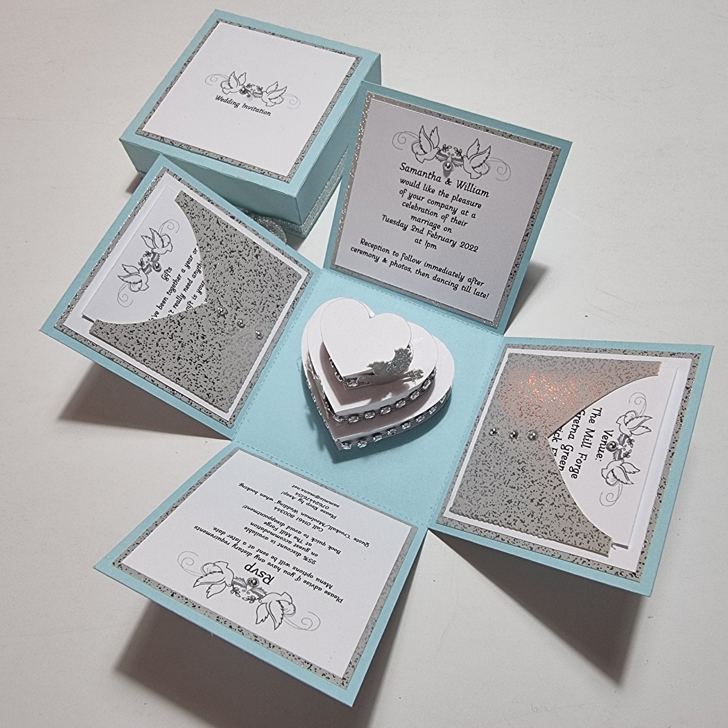 Pastel Blue & Silver Sparkles Exploding Wedding Invitation Box featuring heart-shaped cutouts, a three-tier heart-shaped cake with doves, and intricate invitation details set within a unique design.