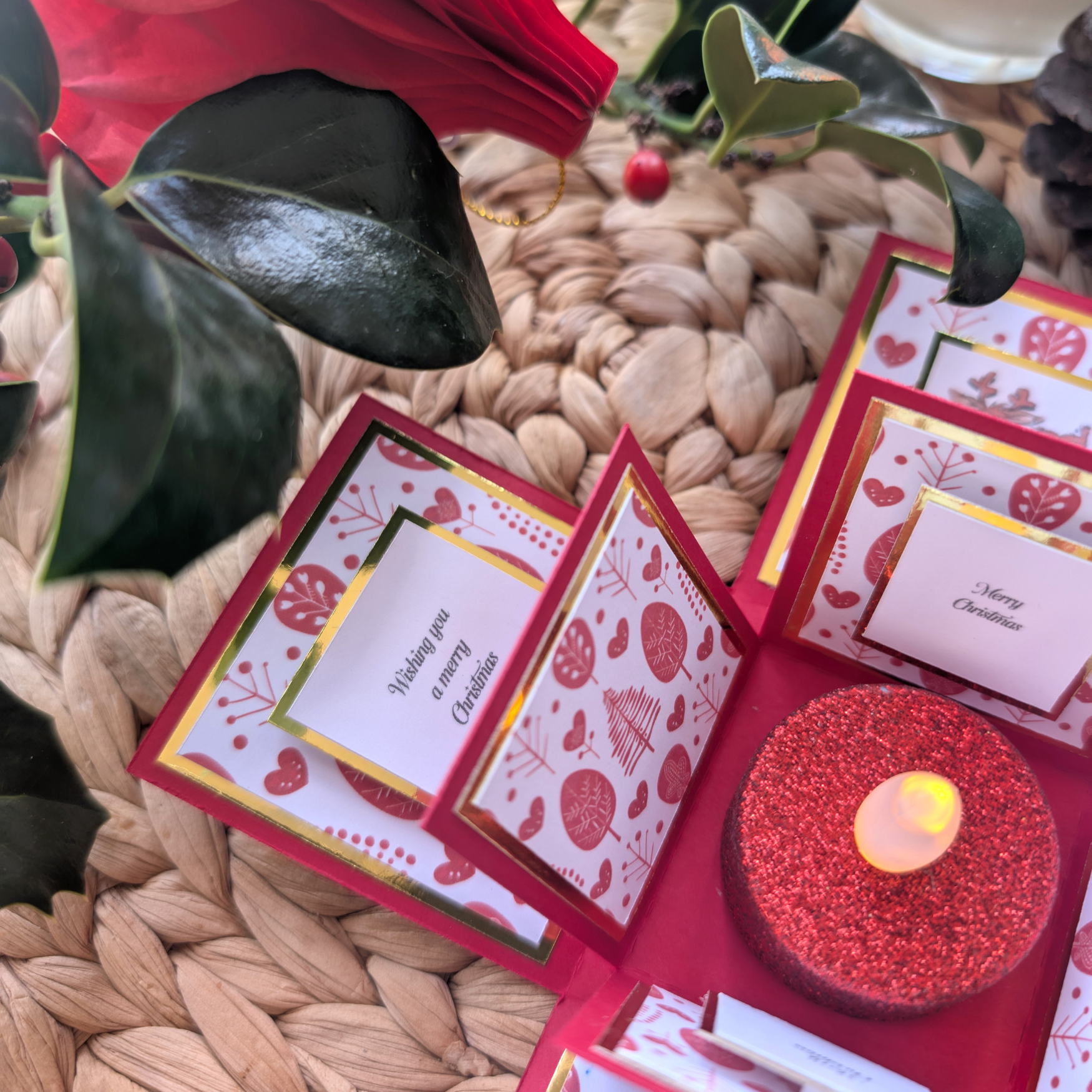 Rudy the Reindeer Exploding Christmas Box Card: a festive pop-up with gold-foiled panels, small greeting cards, and a central red-glittered LED tealight, surrounded by holly leaves, berries, and a pinecone.