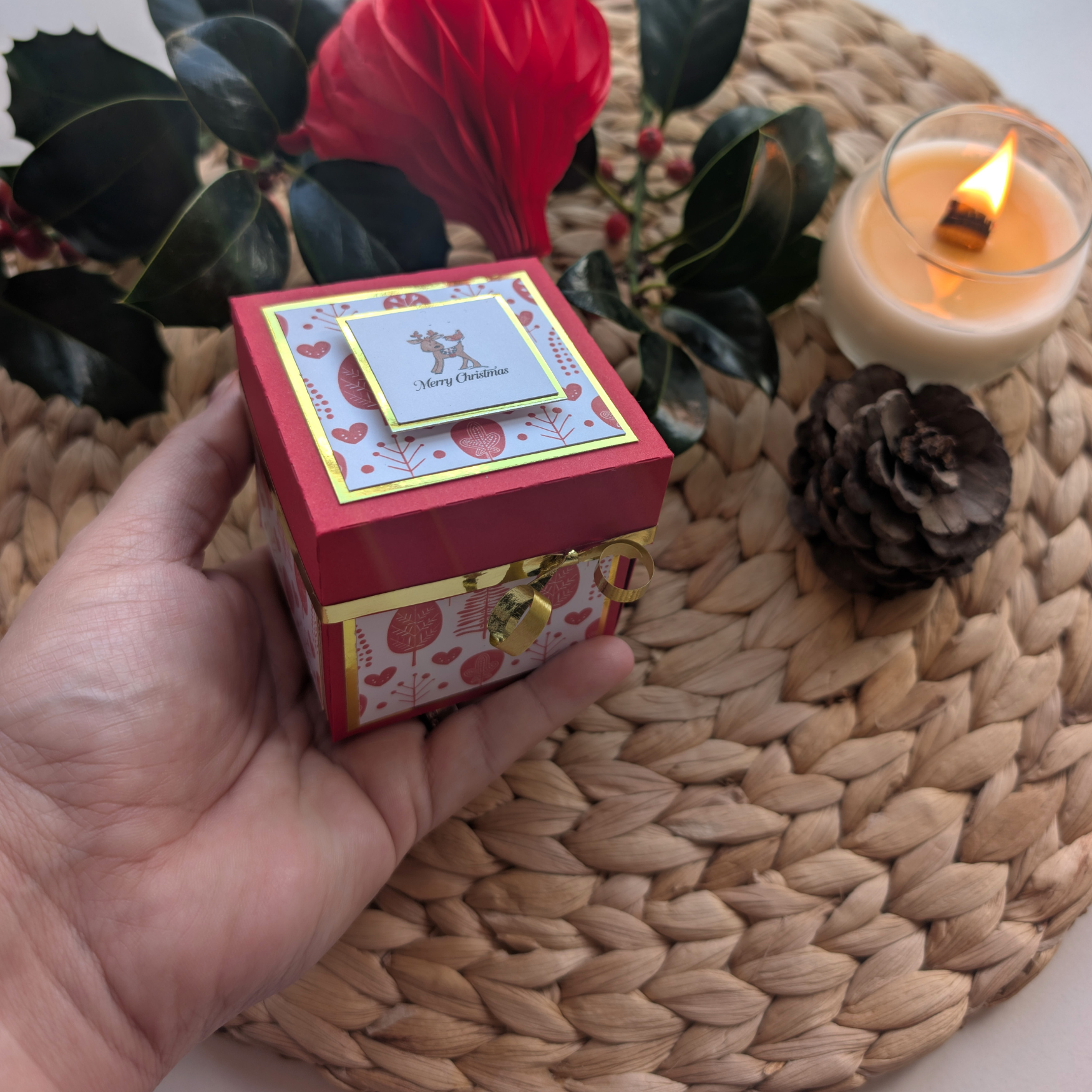 Rudy the Reindeer Exploding Christmas Box Card, a festive red-and-white gift box with gold trim and a reindeer card, is displayed amidst a holiday vignette featuring a candle, pinecone, and greenery.