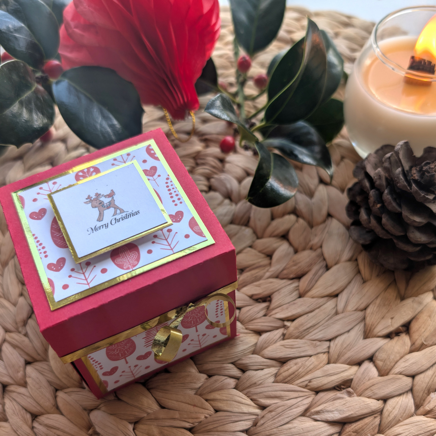 Rudy the Reindeer Exploding Christmas Box Card features a red box with Merry Christmas panel, surrounded by holly, pine cone, and candle, creating a festive, cozy atmosphere.