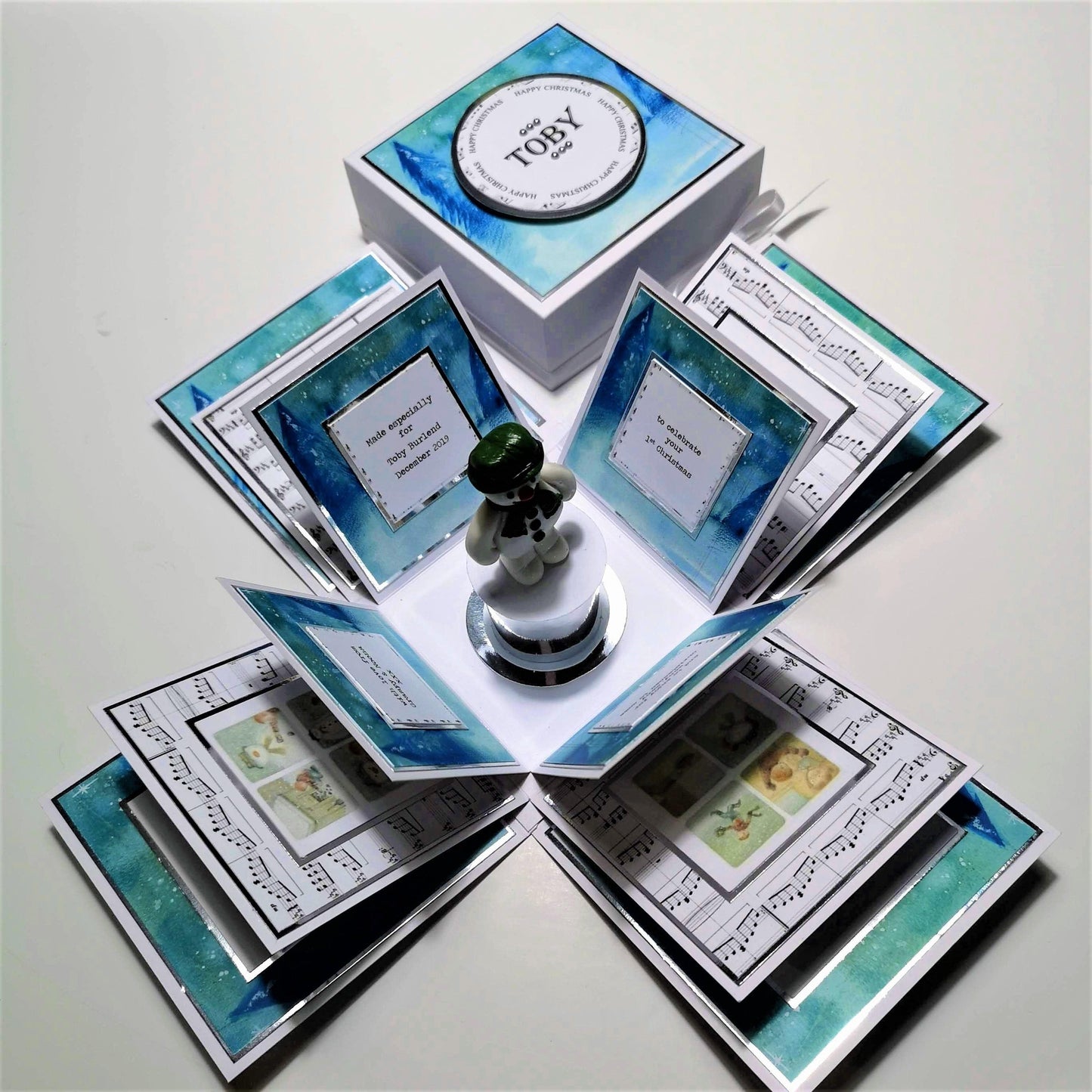 The Snowman Story Box features a white square box with silver accents, containing a snowman statue on a plinth, perfect for a personalized Christmas keepsake.