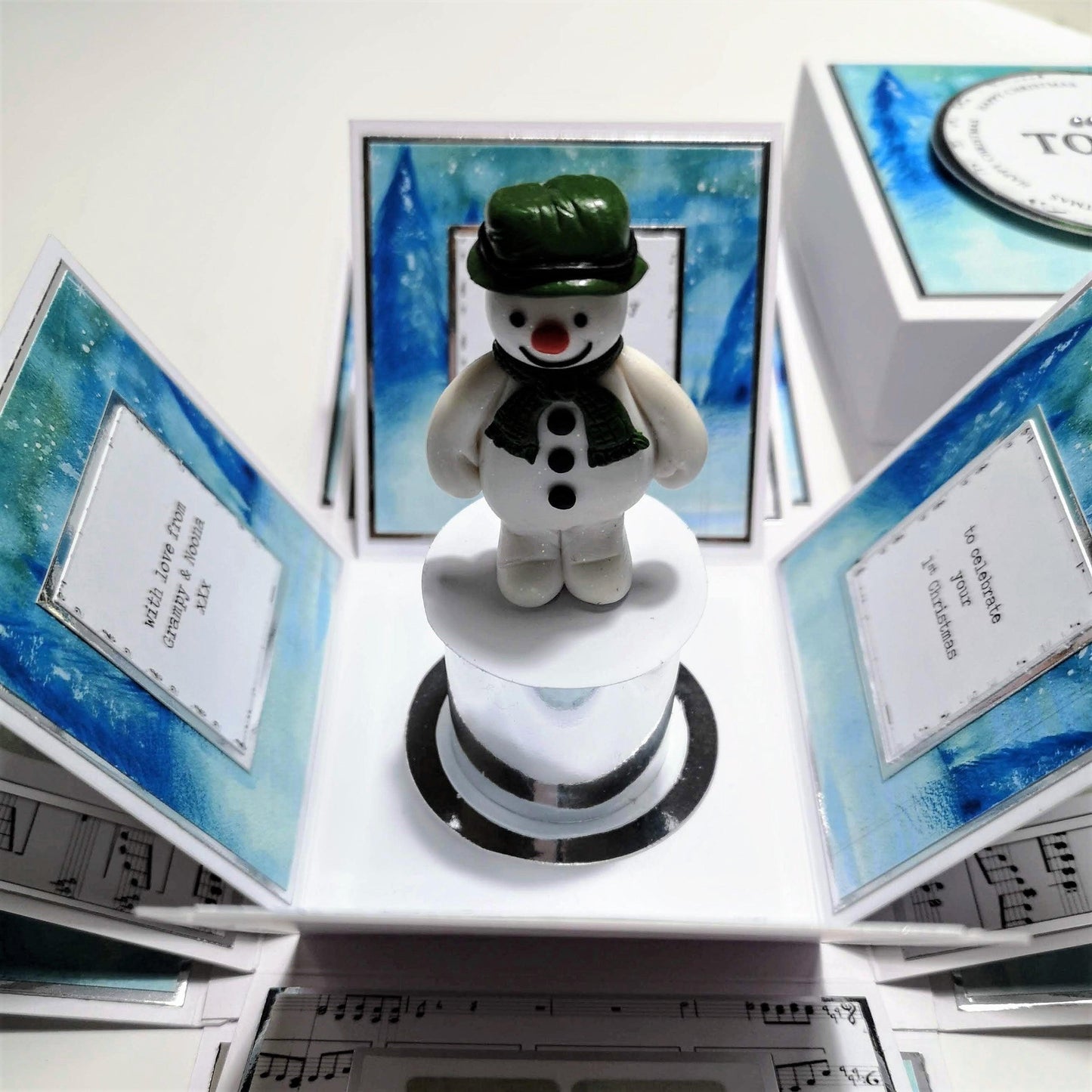 Snowman Story Box featuring a snowman figurine on a white stand, set atop a square white box with silver accents. A unique Christmas keepsake personalized with a name on the lid.