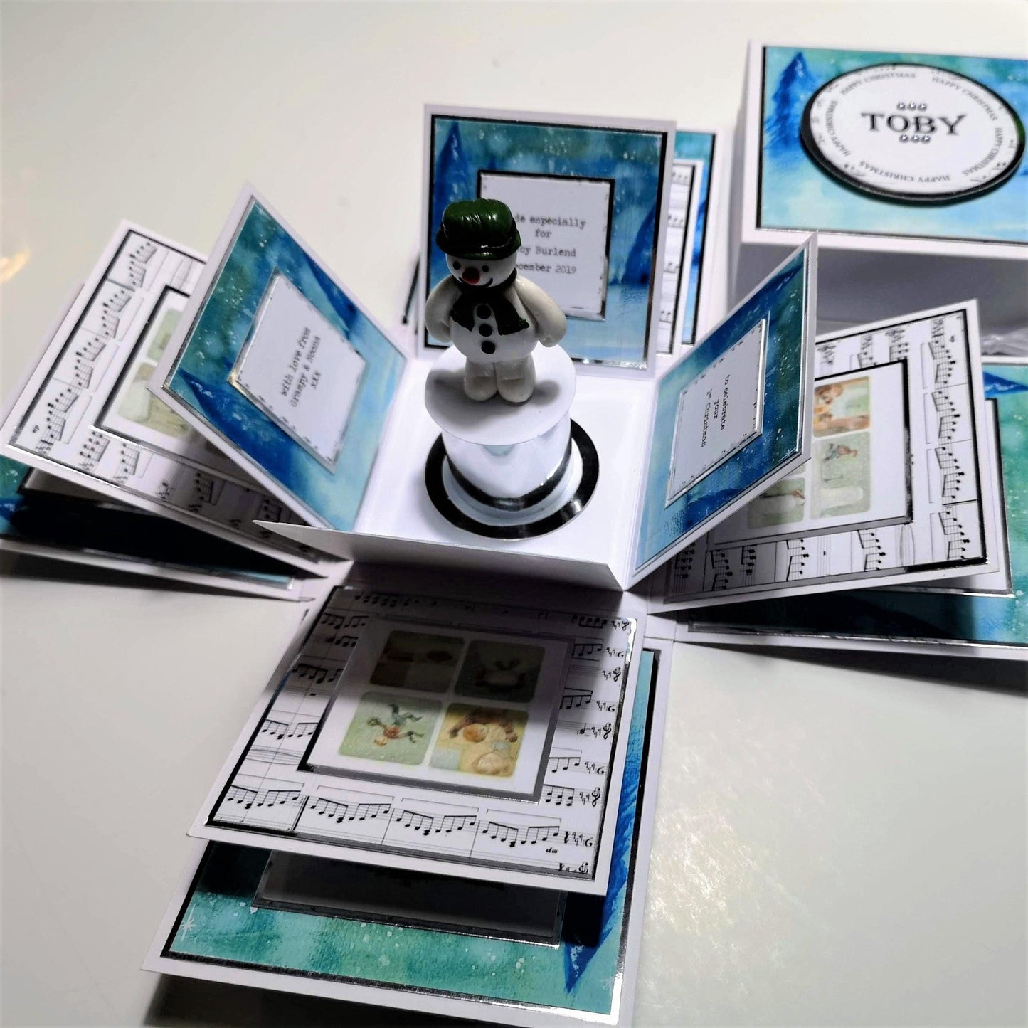 The Snowman Story Box features a snowman figurine on a white plinth inside a 4'' square white cardstock box, complemented by silver accents and personalized with a name on the lid.