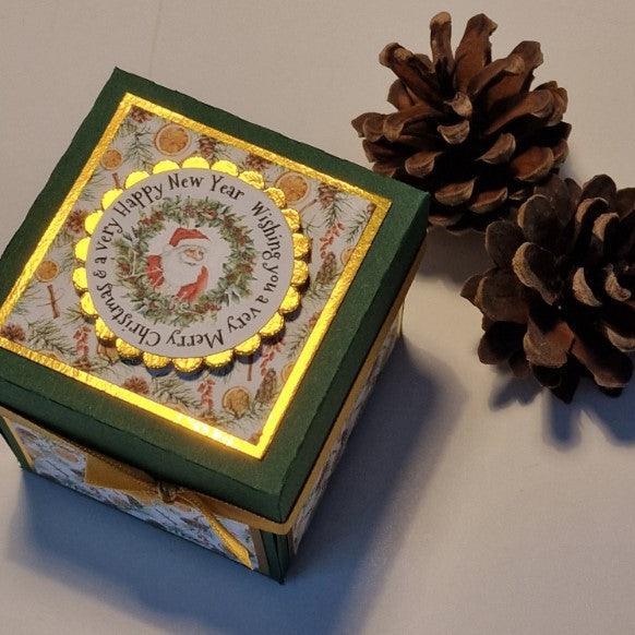 Vintage styled Christmas Candle Box: A green box with a gold ribbon and bow, adorned with festive decorations, next to pine cones. Features a Santa Claus plate, perfect for holiday gift-giving.