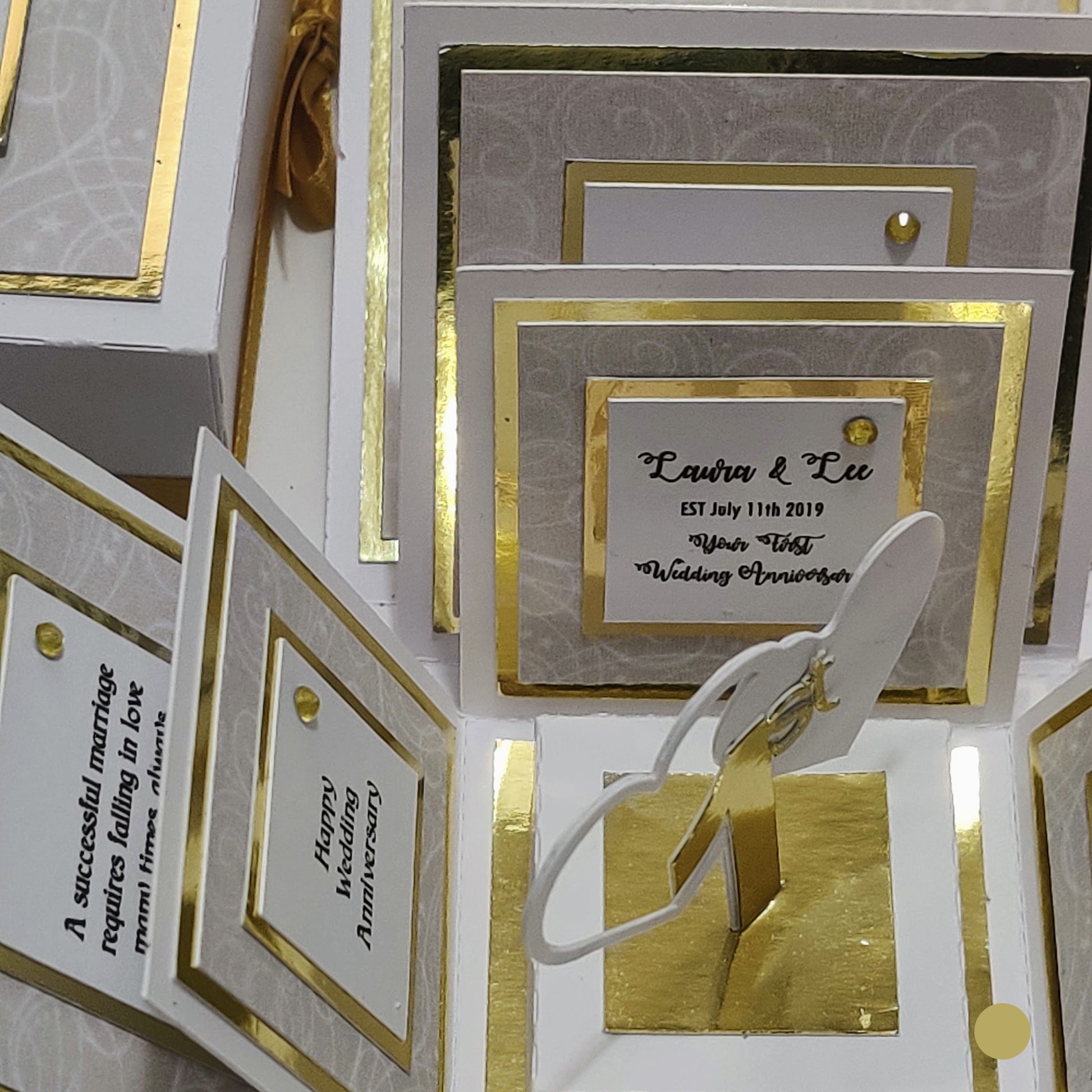 First Wedding Anniversary Exploding Box, a 3-D pop-up keepsake card with white panels edged in gold, features personalized wedding-anniversary messages and a central gold stand for a photo or decoration.