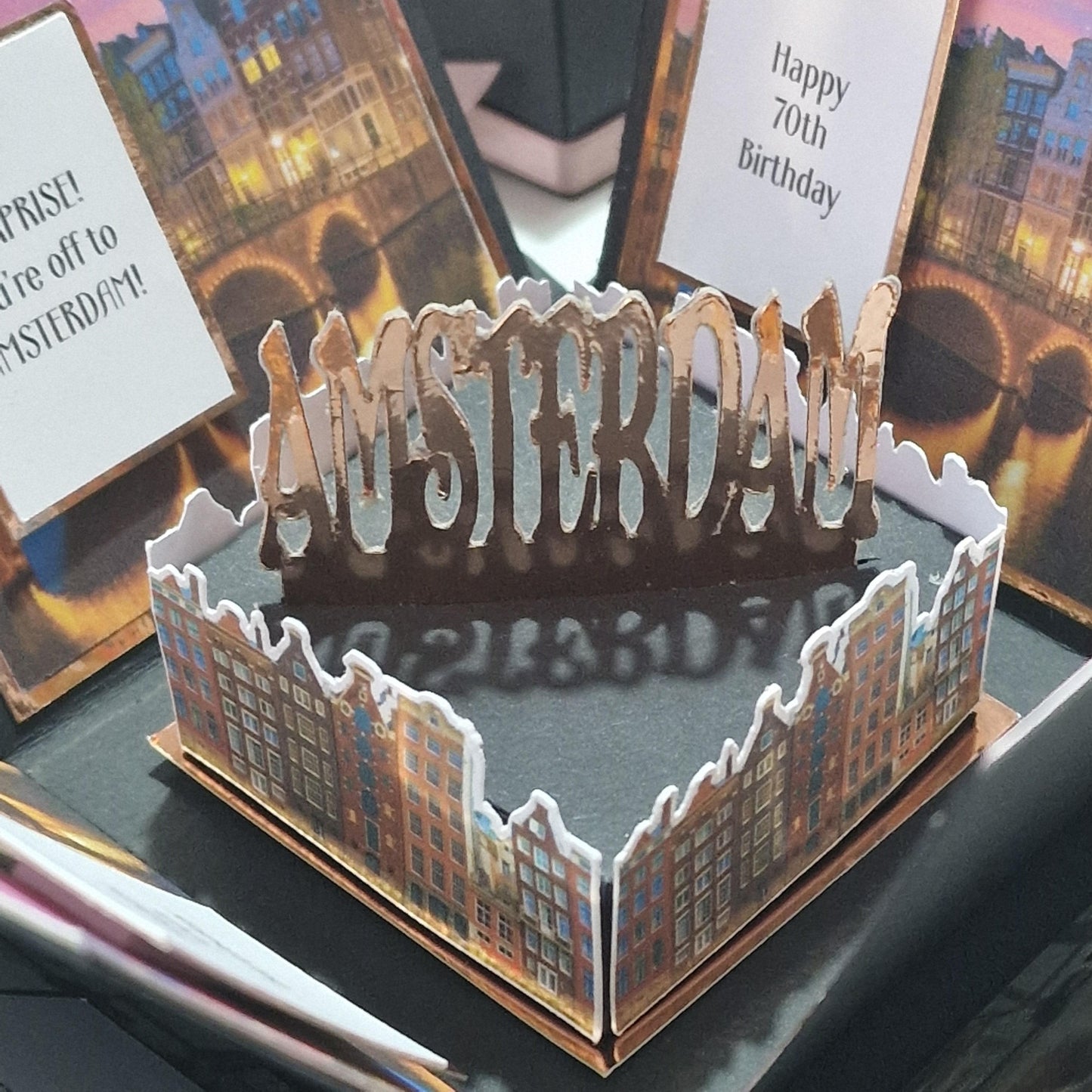 Exploding Occasions Box - Trip Reveal AMSTERDAM featuring a 3D Amsterdam cityscape centerpiece, surrounded by decorative paper panels and event details, perfect for a unique birthday or Christmas surprise.