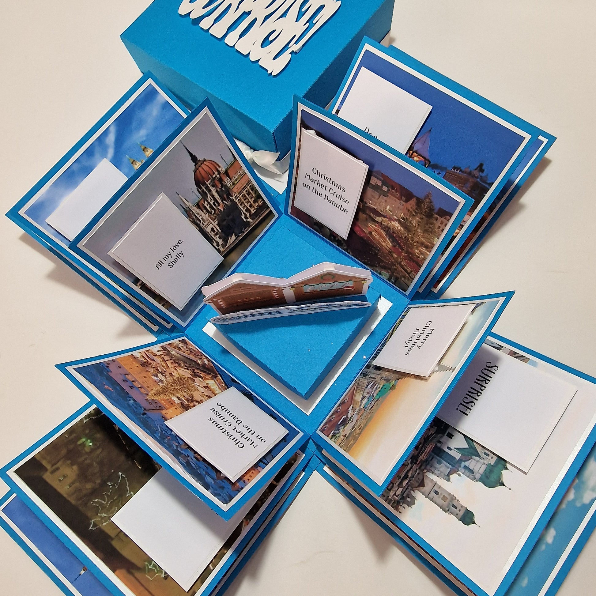 Christmas Markets Exploding Trip Reveal Gift Box featuring a blue box with white text, designed for holiday surprise, incorporating a festive Christmas tree and wooden-style chalets inside for a unique gifting experience.