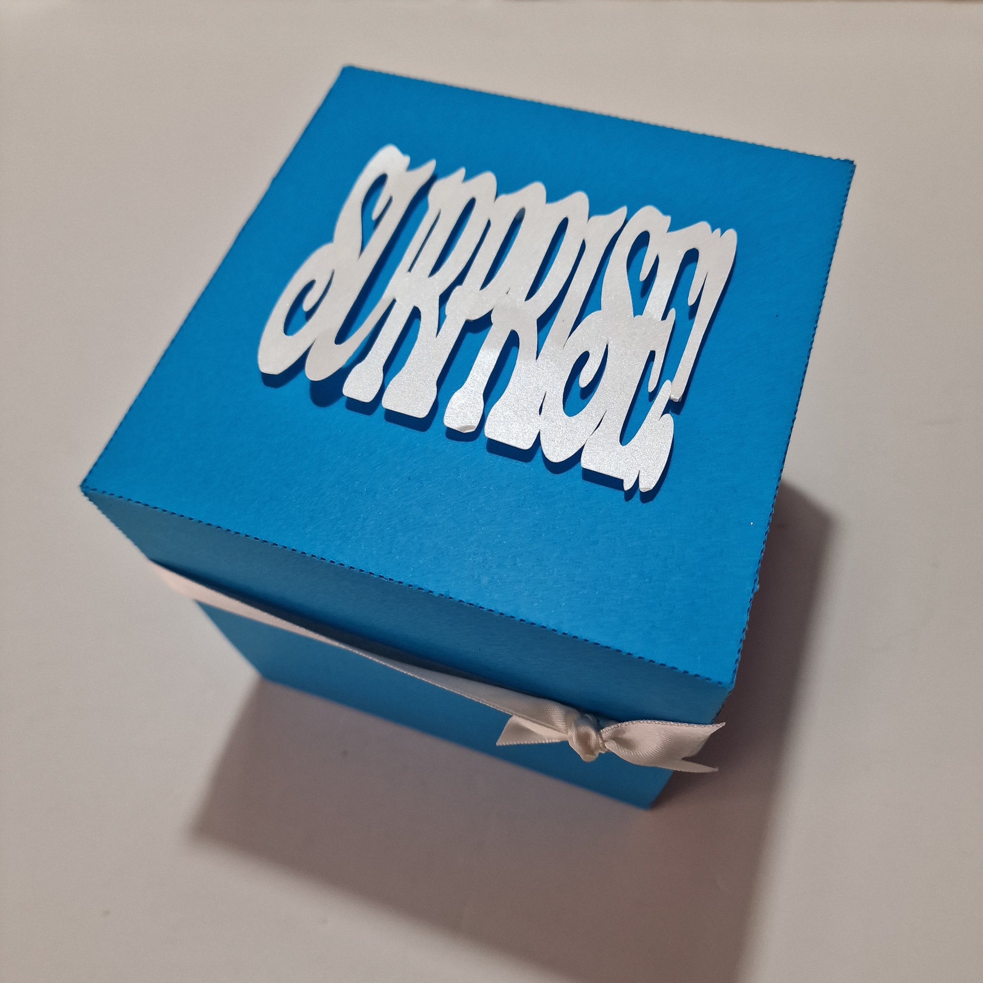 Christmas Markets Exploding Trip Reveal Gift Box featuring intricate white text on a blue box, designed to unveil holiday plans with a charming Christmas tree and wooden-style chalets inside.