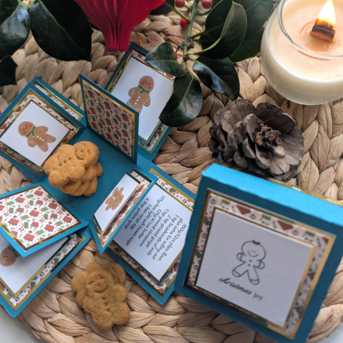 Exploding Christmas Box Card with Gingerbread Recipe showcasing festive panels with gingerbread-man illustrations and real mini cookies. Surrounded by holiday decor like a candle, pine cone, and holly for a cozy presentation.