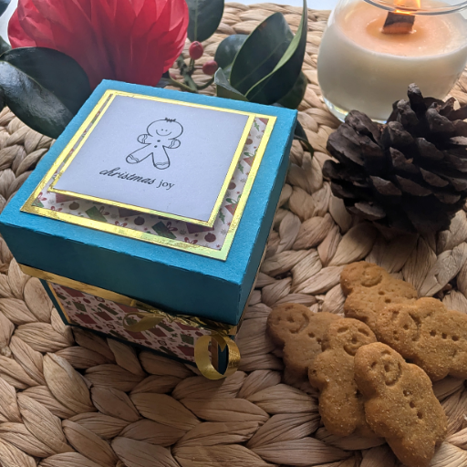 Exploding Christmas Box Card with Gingerbread Recipe features a teal lid, holly-patterned sides, and a gingerbread-man card. Accompanied by cookies, candle, pine cone, and greenery on a woven tray.