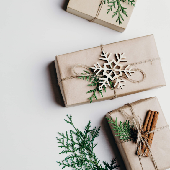 Three gift boxes wrapped in brown paper with string and decorative snowflake, surrounded by greenery on a light background.