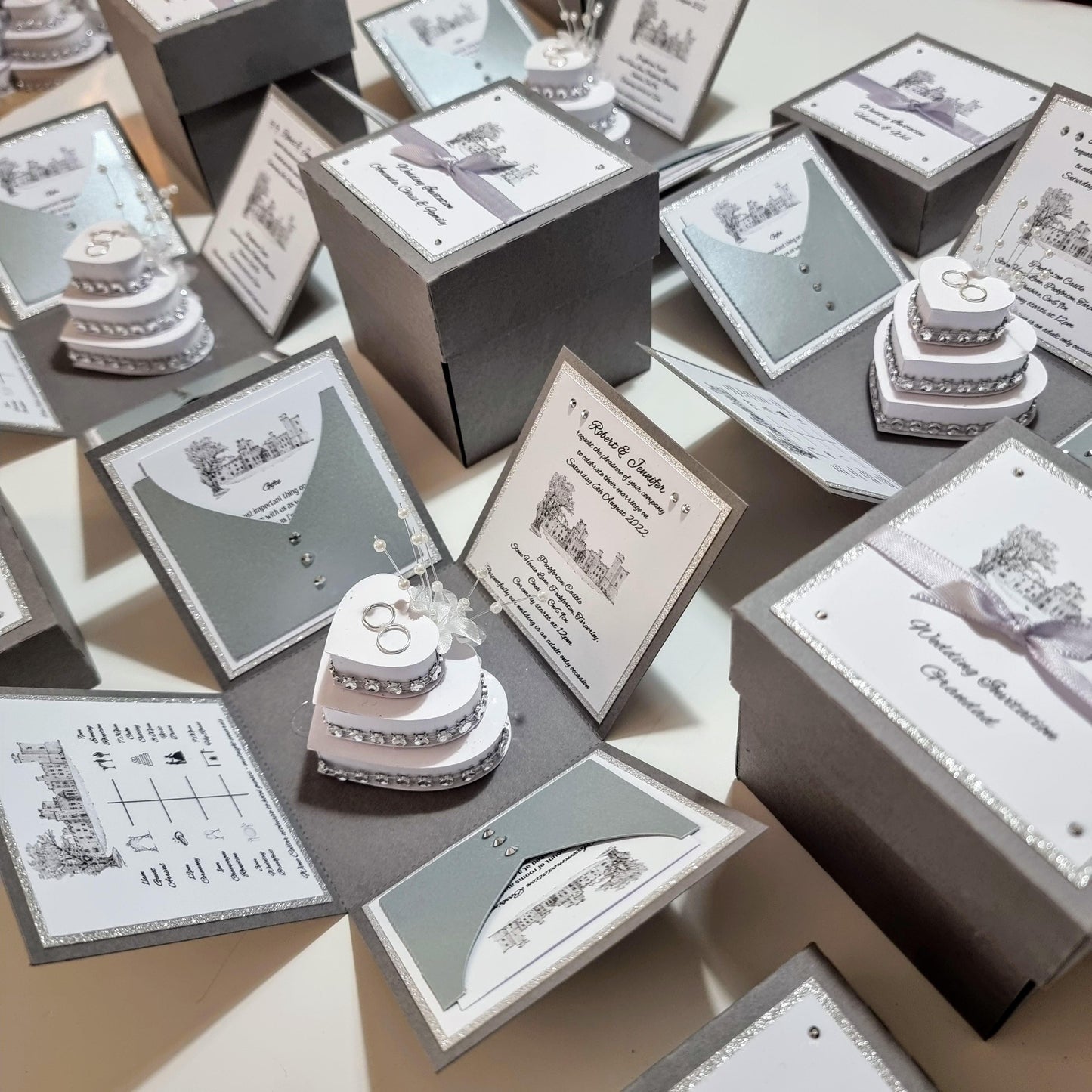 Illustrated Style Exploding Wedding Invitation Boxes featuring a unique square design, with panels for invitations and event details, and a heart-shaped cake centerpiece adorned with rings, perfect for luxury wedding stationery.