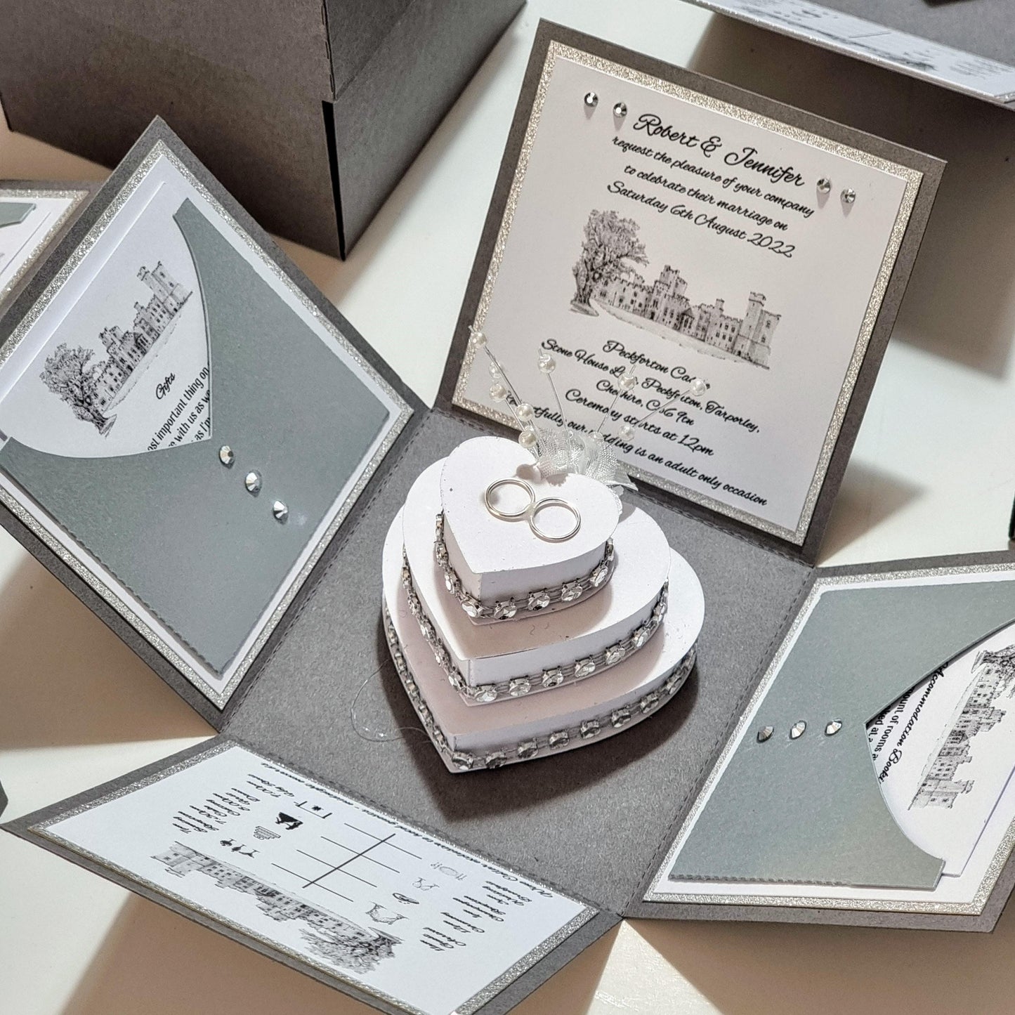 Illustrated Styled Exploding Wedding Invitation Box featuring a heart-shaped cake with rings, venue illustration, and interactive panels for event details, menu choices, and RSVP cards.