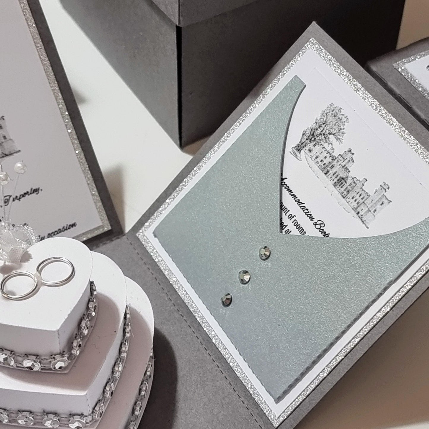 Illustrated Styled Exploding Wedding Invitation Boxes featuring a silver card with a castle illustration, silver envelope, and a heart-shaped cake topped with rings, embodying luxury invites and bespoke wedding stationery.