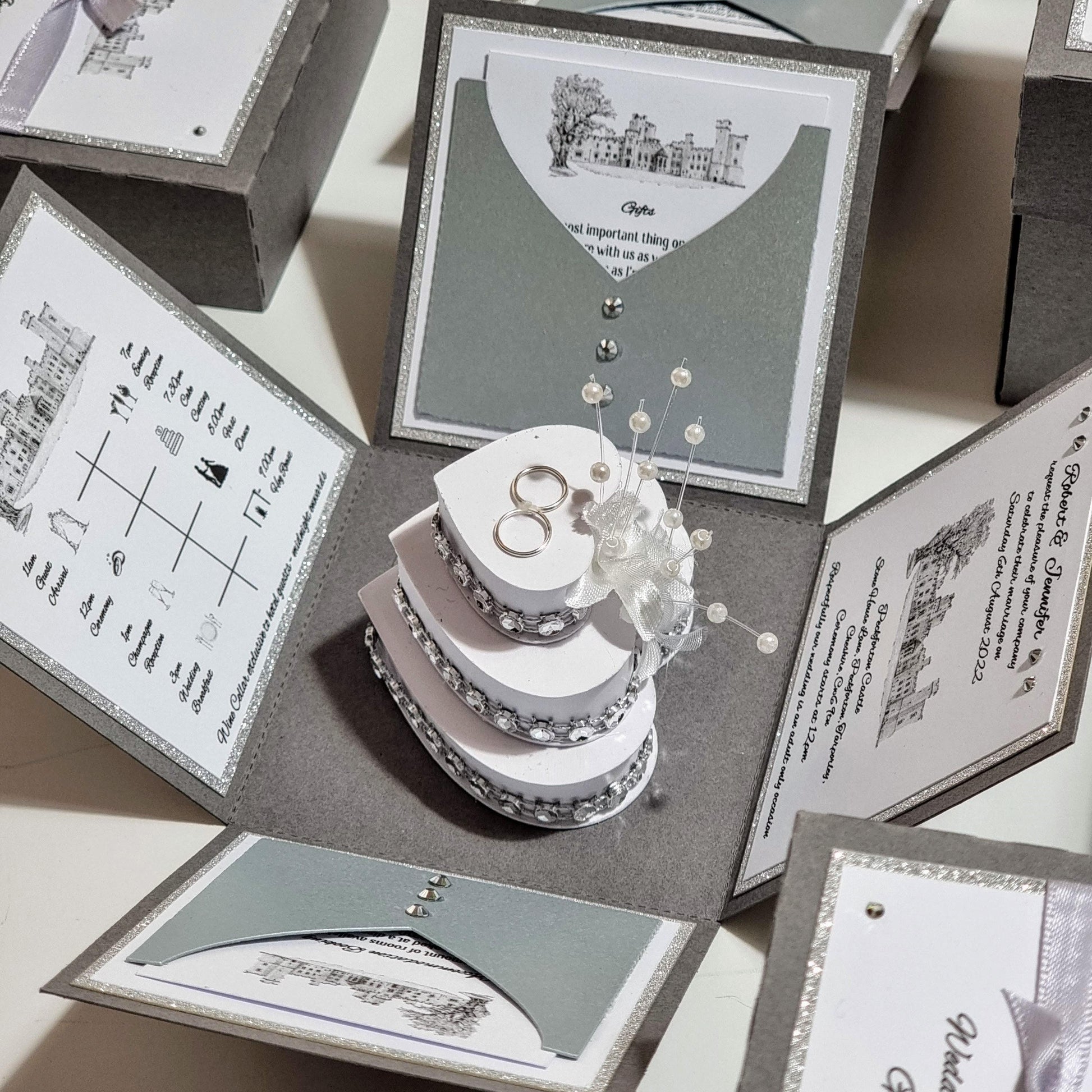 Illustrated Style Exploding Wedding Invitation Box showcasing a heart-shaped cake with silver rings and an illustrated venue card, designed with luxury invites and bespoke stationery elements.