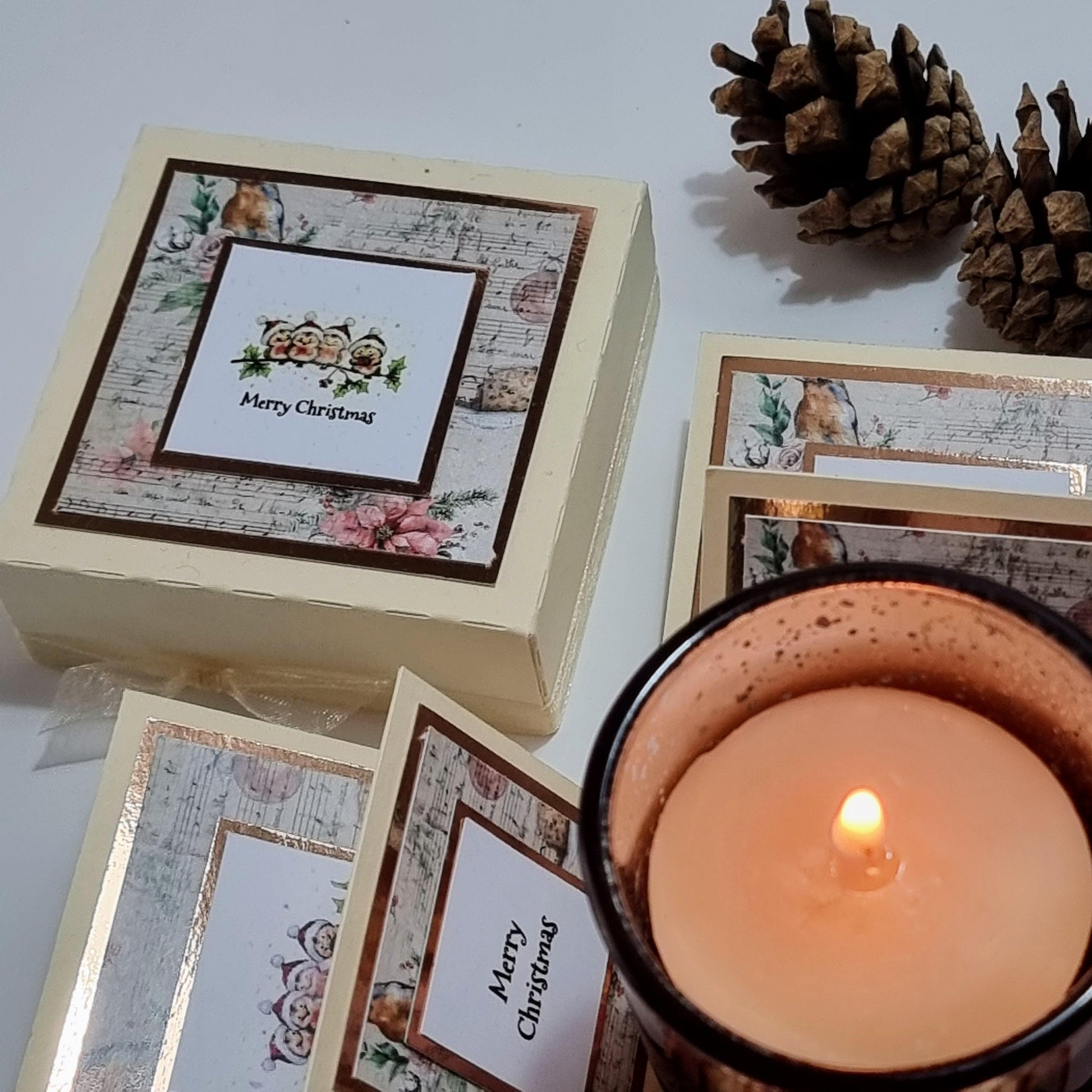 Rocking Robin Christmas Candle Box featuring a glass votive candleholder and pine cones on a table, with a square card showing Christmas robins and a festive greeting.