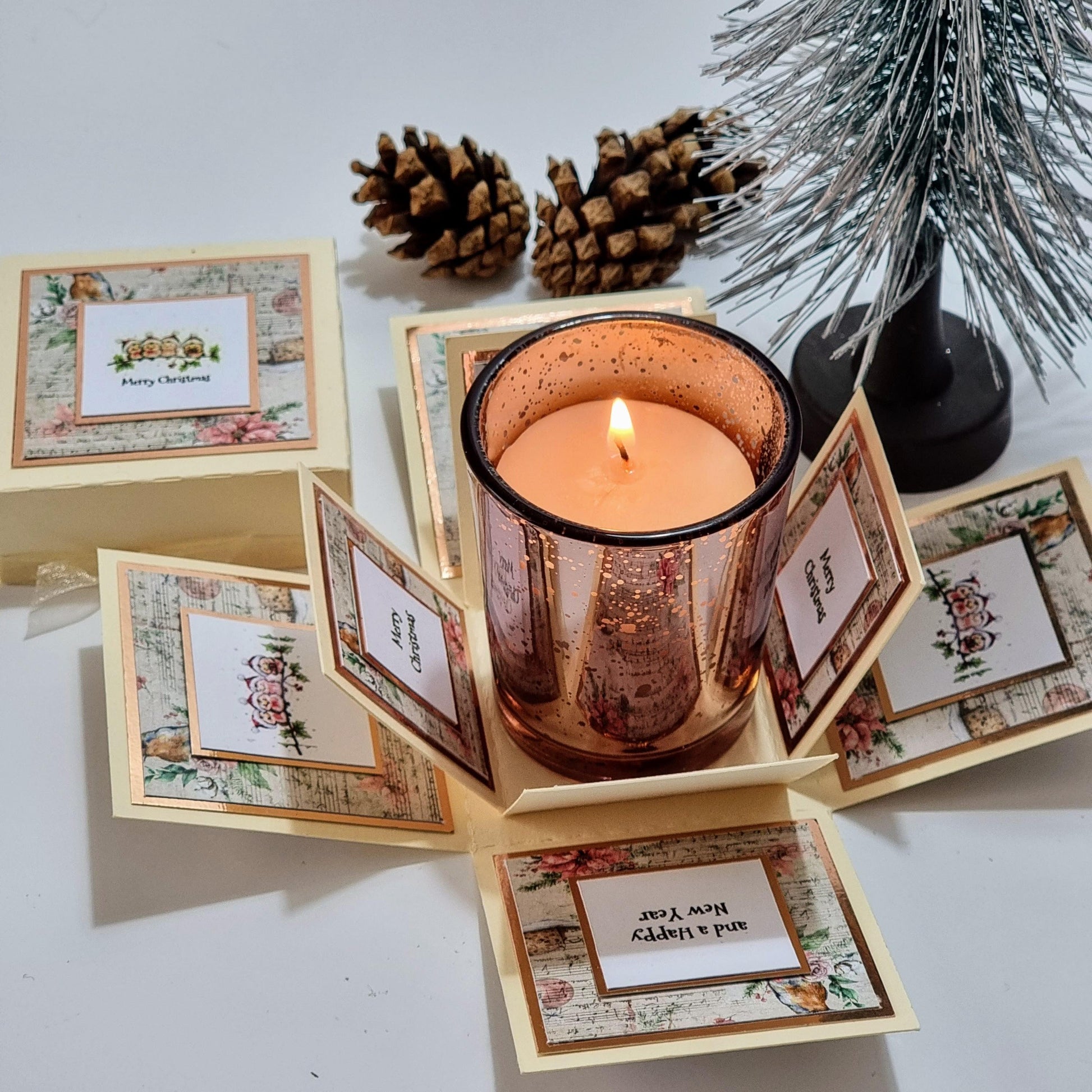 Rocking Robin Christmas Candle Box featuring a votive candleholder with Christmas-scented candle, surrounded by pine cones and a small tree, set in a clotted cream box with rose gold accents.