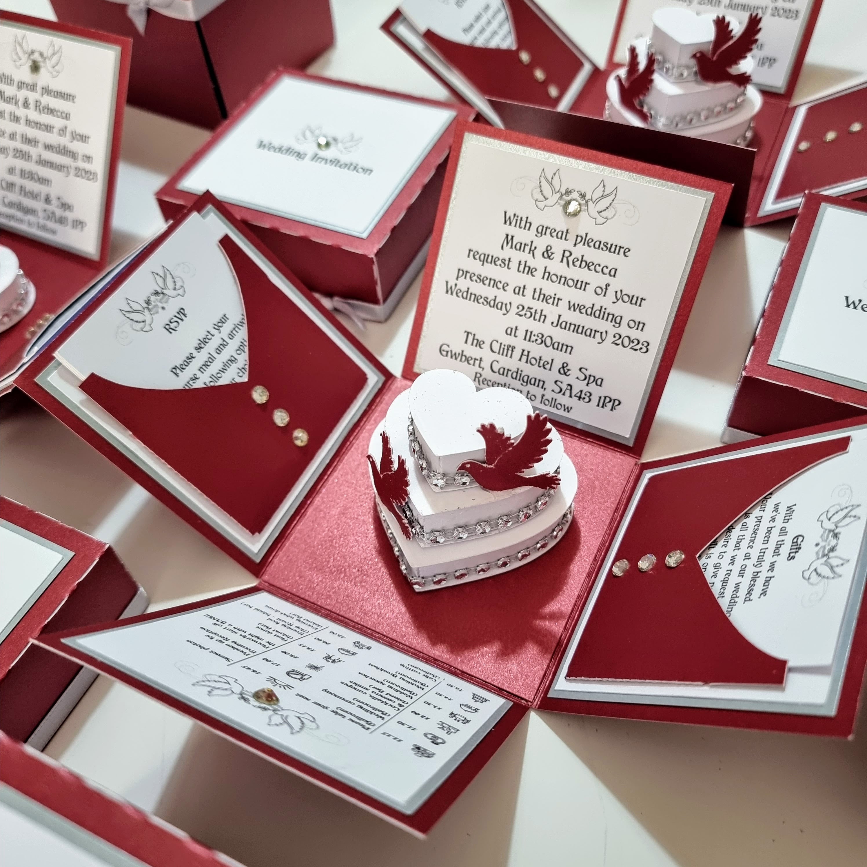 The image shows a collection of intricately designed wedding invitations. They are red and white with a pop-up heart and bird design in the center. The text on the main invitation reads:

"With great pleasure, Mark & Rebecca request the honor of your presence at their wedding on Wednesday 25th January 2023 at 11:30 am. The Cliff Hotel & Spa Gwbert, Cardigan, SA43 1PP. Reception to follow."

The invitations also include RSVP cards and other details about the wedding. The design is elegant and detailed