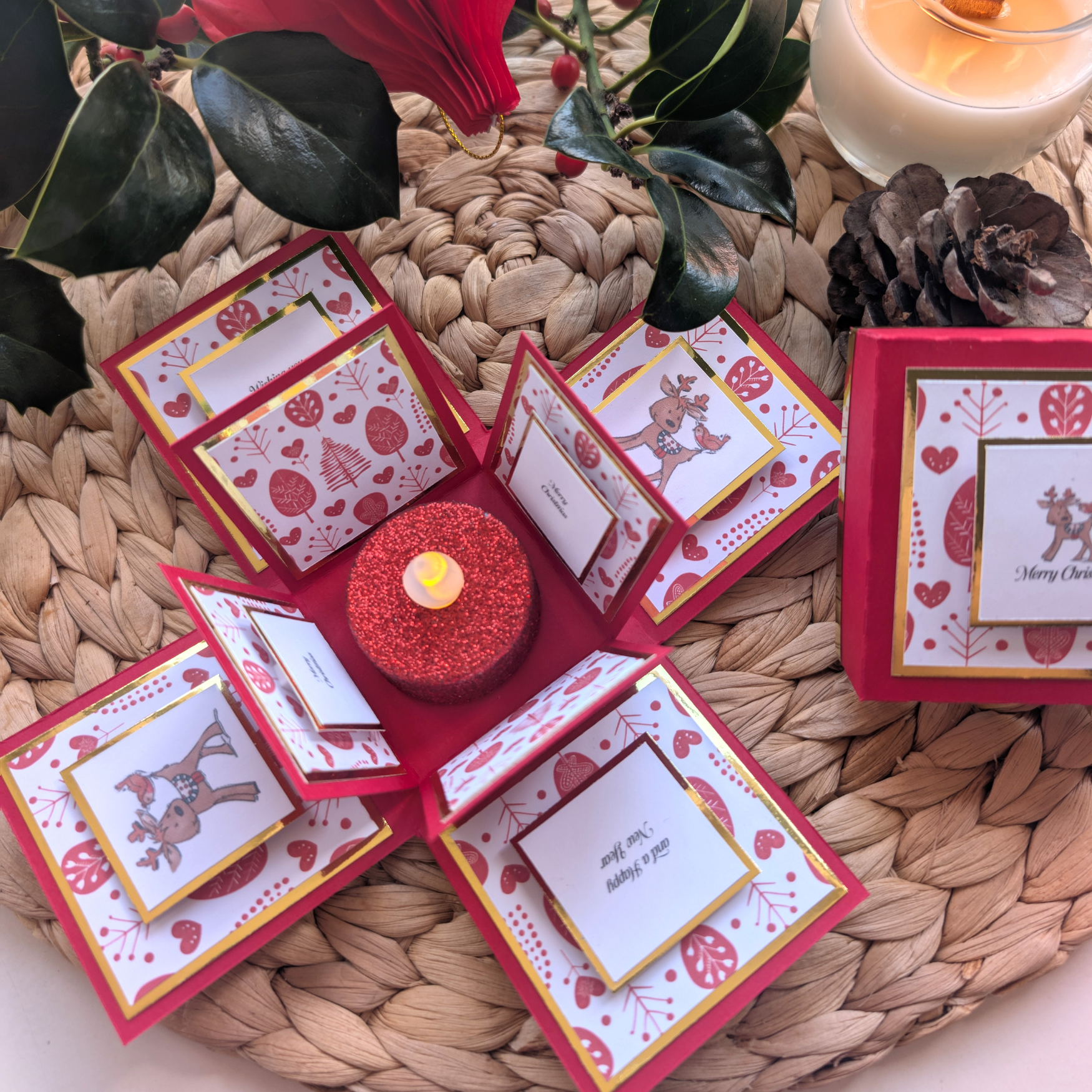 Rudy the Reindeer Exploding Christmas Box Card: A pop-up card with festive panels featuring reindeer, hearts, and trees, centered around a red glittered LED tea light, perfect for spreading holiday cheer.