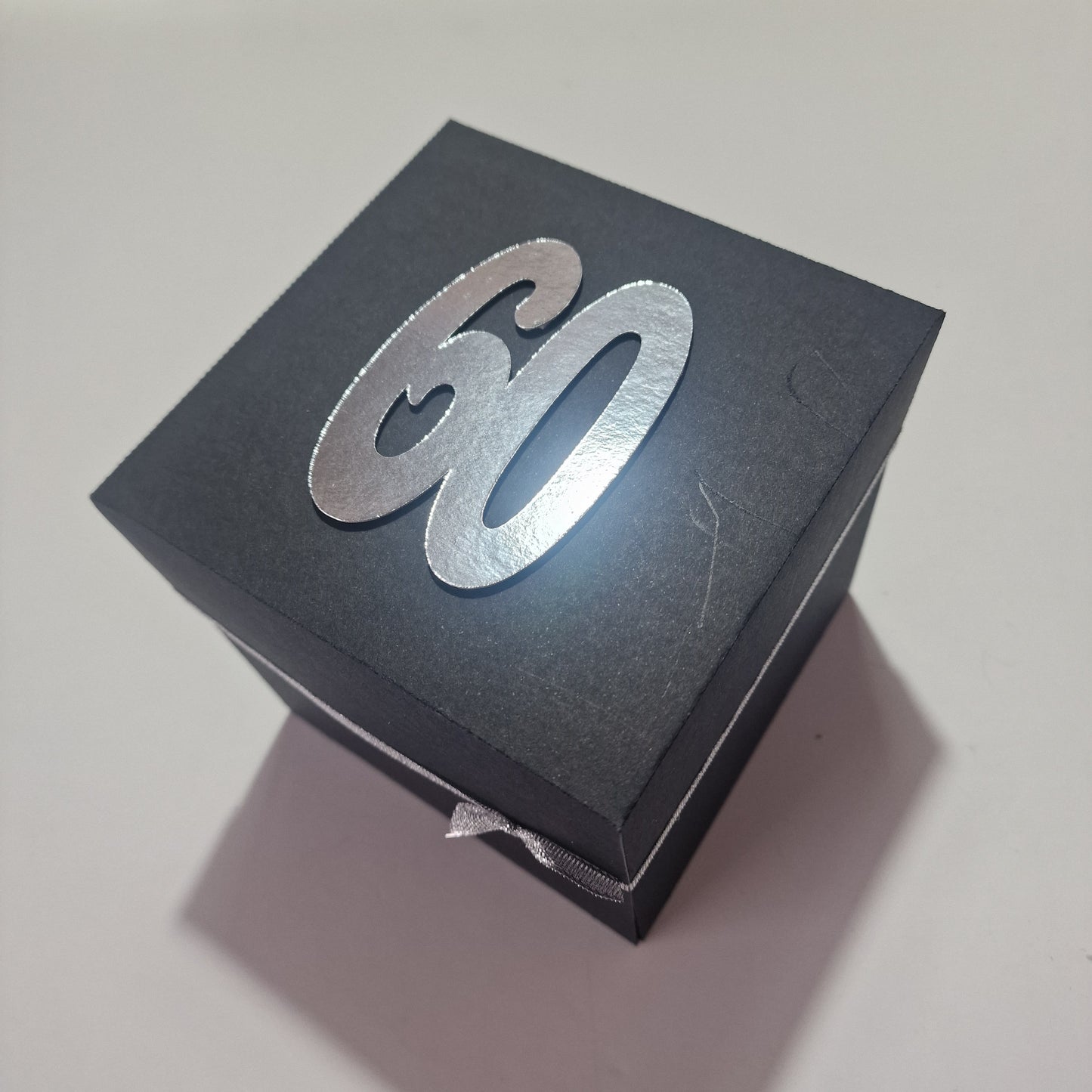 Santorini Exploding Trip Reveal Box features a matte-black, square design with a silver 60 on the lid, hinting at a surprise trip to Santorini with personalized layers inside.