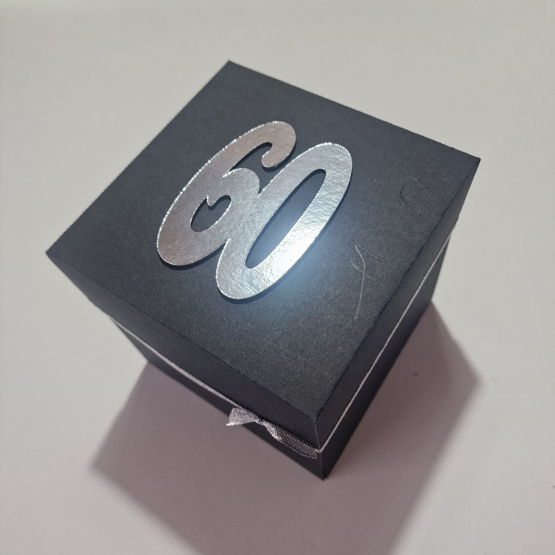 Santorini Exploding Trip Reveal Box features a matte-black, square design with a silver 60 on the lid, hinting at a surprise trip to Santorini with personalized layers inside.