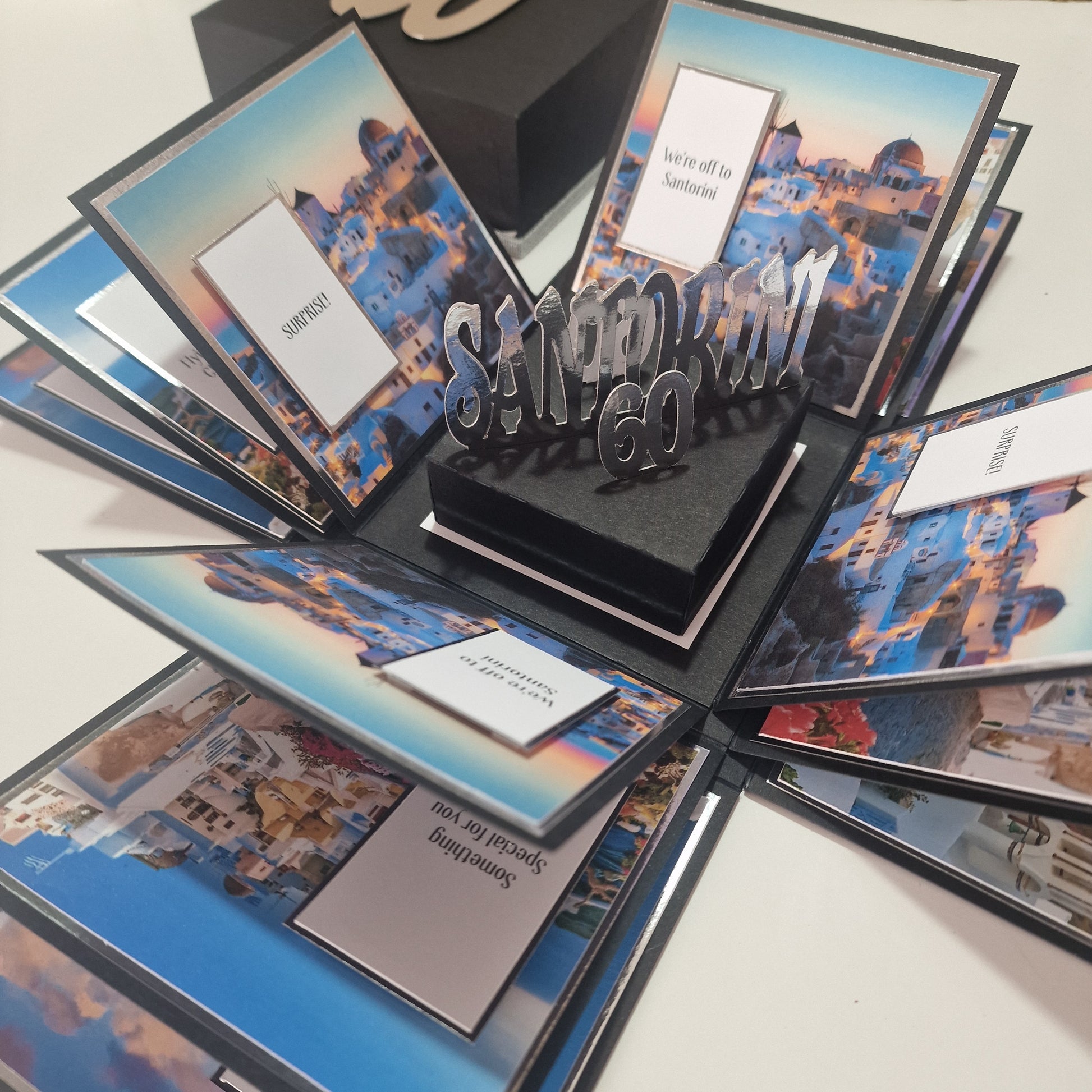 Santorini Exploding Trip Reveal Box: A pop-up gift box featuring Santorini-themed photos and messages, opening to a SANTORINI GO pop-up, creatively announcing a Santorini trip.