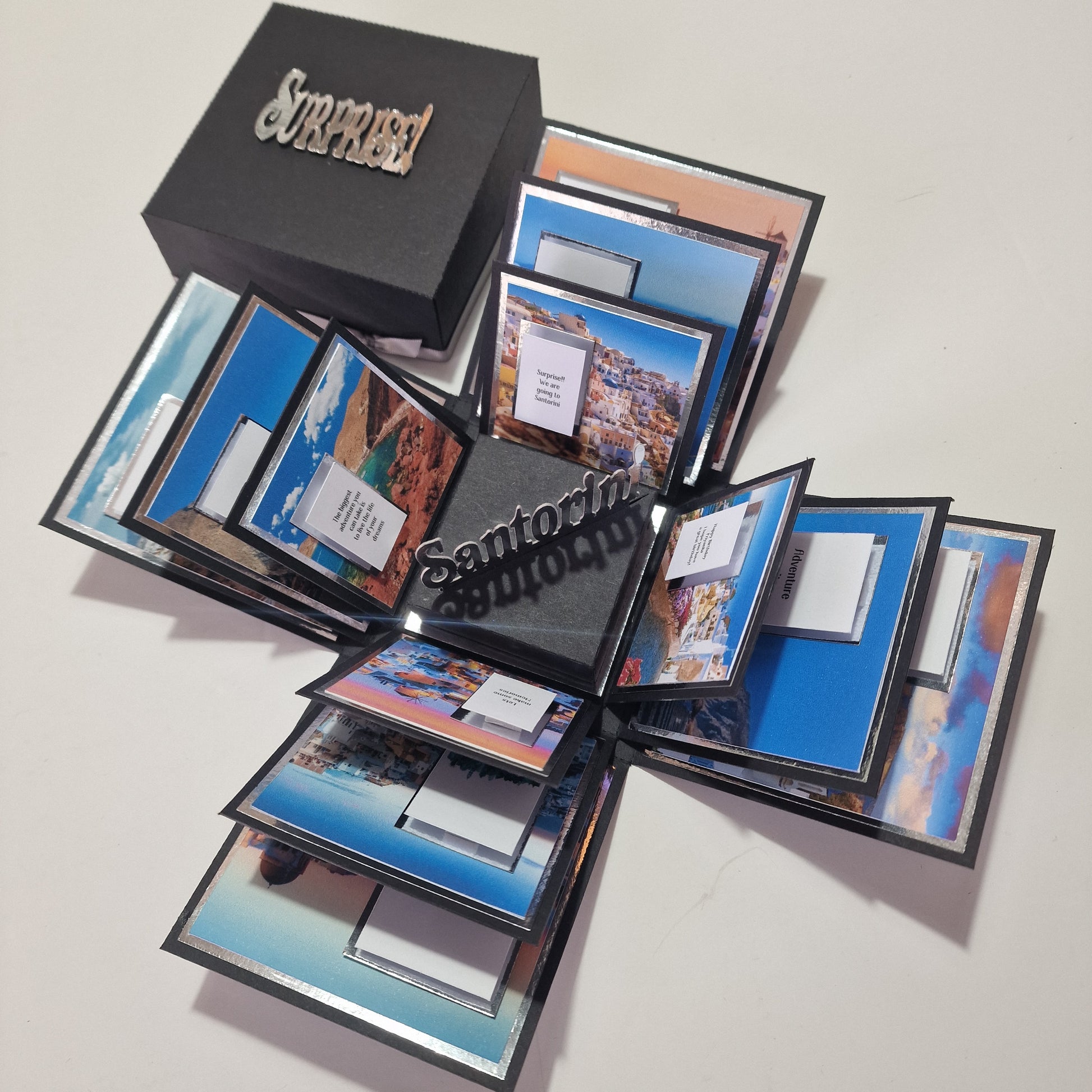 Santorini Exploding Trip Reveal Box – Unique Holiday Surprise Gift, features opened panels with Santorini photos and note cards, lid reading “Surprise!” and center panel spelling “Santorini,” announcing a memorable trip.
