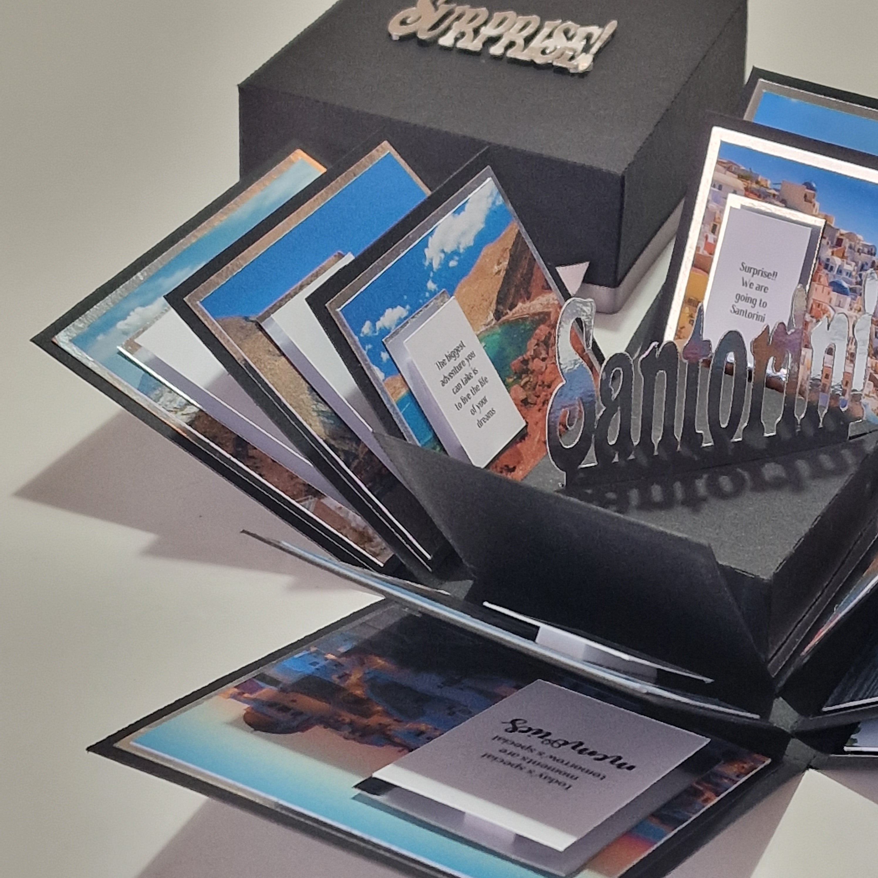 Santorini Exploding Trip Reveal Box features a black cube that unfolds into panels with Santorini vistas and travel messages, revealing a surprise trip. Perfect for an unforgettable holiday announcement.