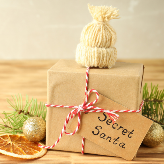 Gift wrapped in brown paper with a 'Secret Santa' tag, topped with a knitted hat, on a wooden surface with Christmas decorations.