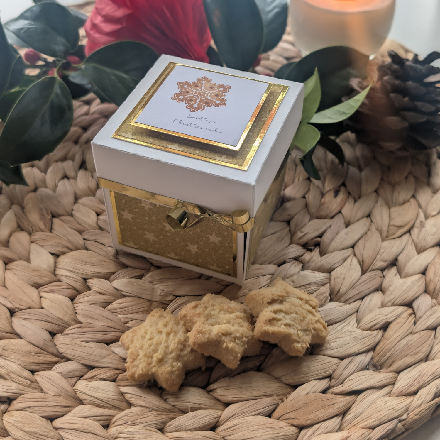 Exploding Christmas Box Card with Shortbread Recipe – Handmade Festive Keepsake: White gift box with gold snowflake motif and cookies on a natural placemat, set amidst evergreen sprigs and a candle.