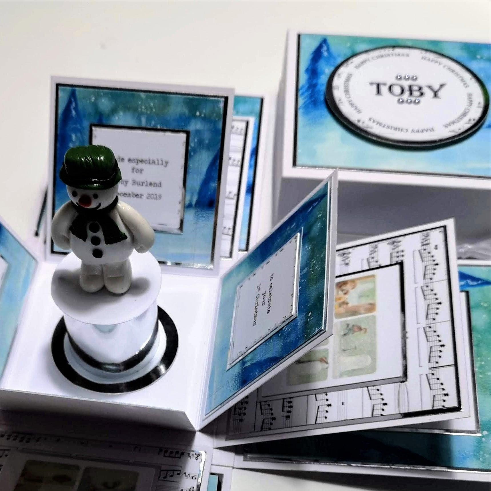 The Snowman Story Box features a snowman figurine on a white box, accompanied by blue and white cards, highlighting its unique Christmas theme and personalized design.
