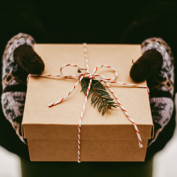 Brown gift box with string and greenery, held by hands in patterned gloves against a dark background