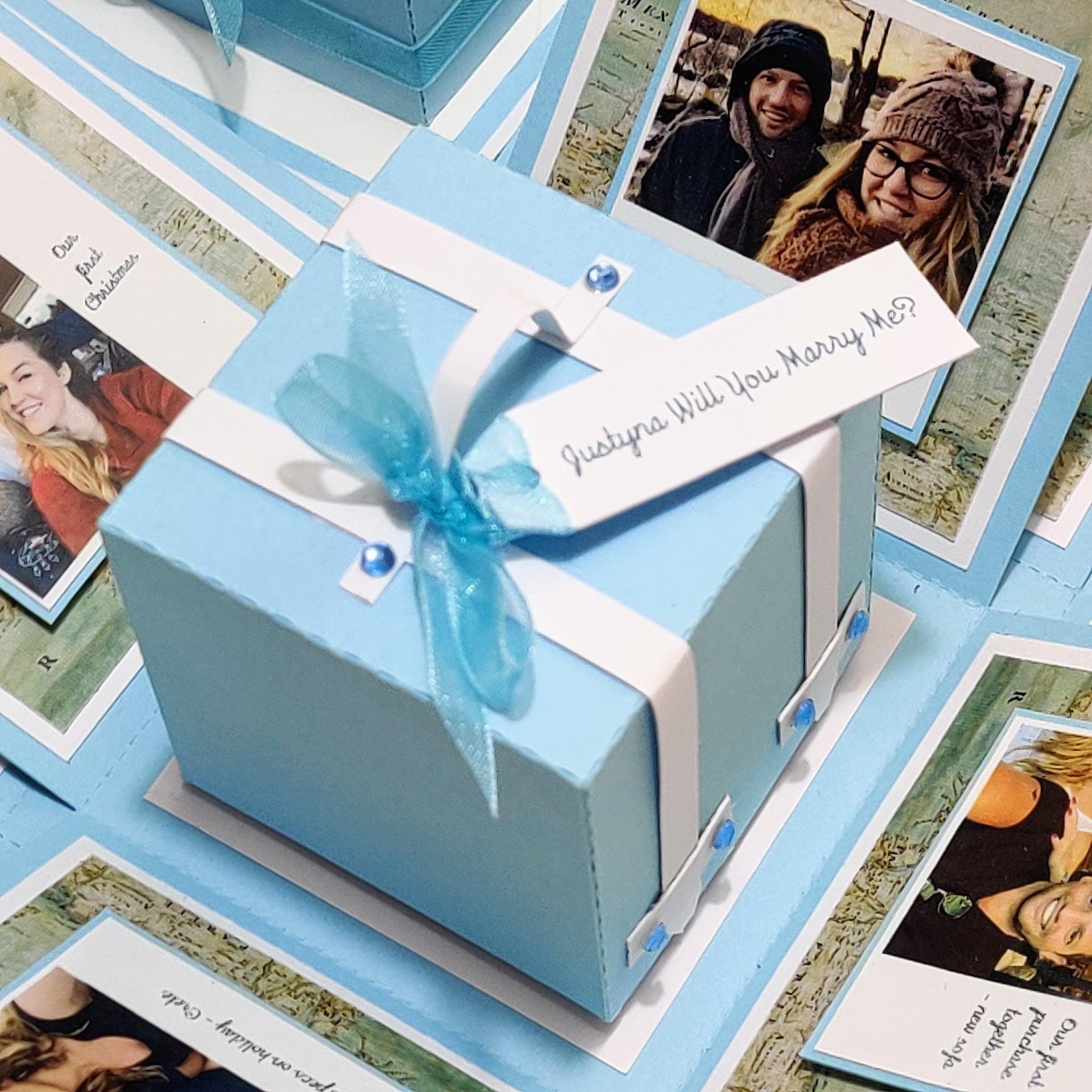 Travel Themed Proposal Box with a blue box adorned with a ribbon and bow, sitting atop a pile of photos, perfect for an unforgettable engagement ring presentation.