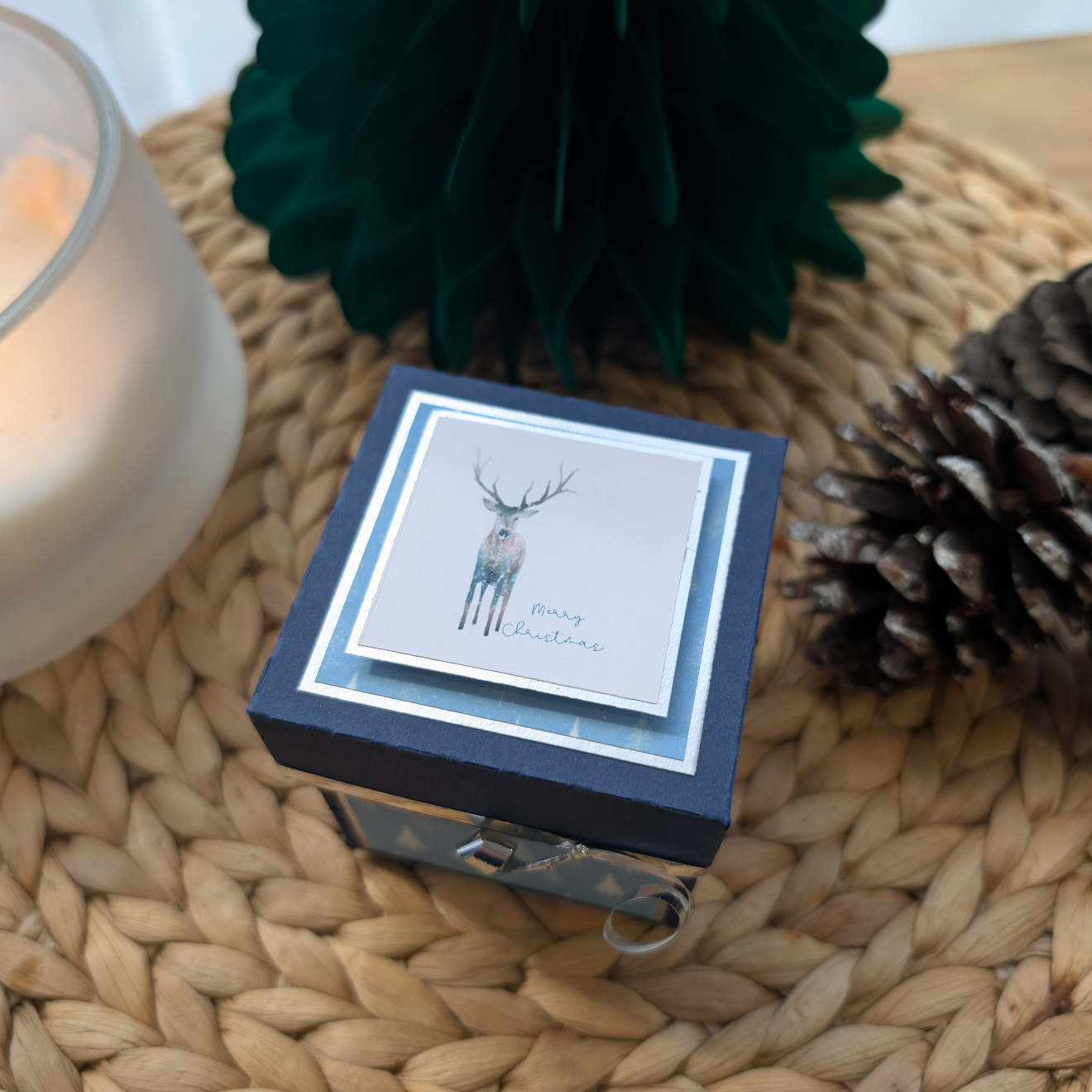 Winter Stagg Christmas Box Card on a placemat, featuring a watercolor stag and Merry Christmas. Cozy with pinecones, a candle, and a green paper tree, capturing festive holiday charm.