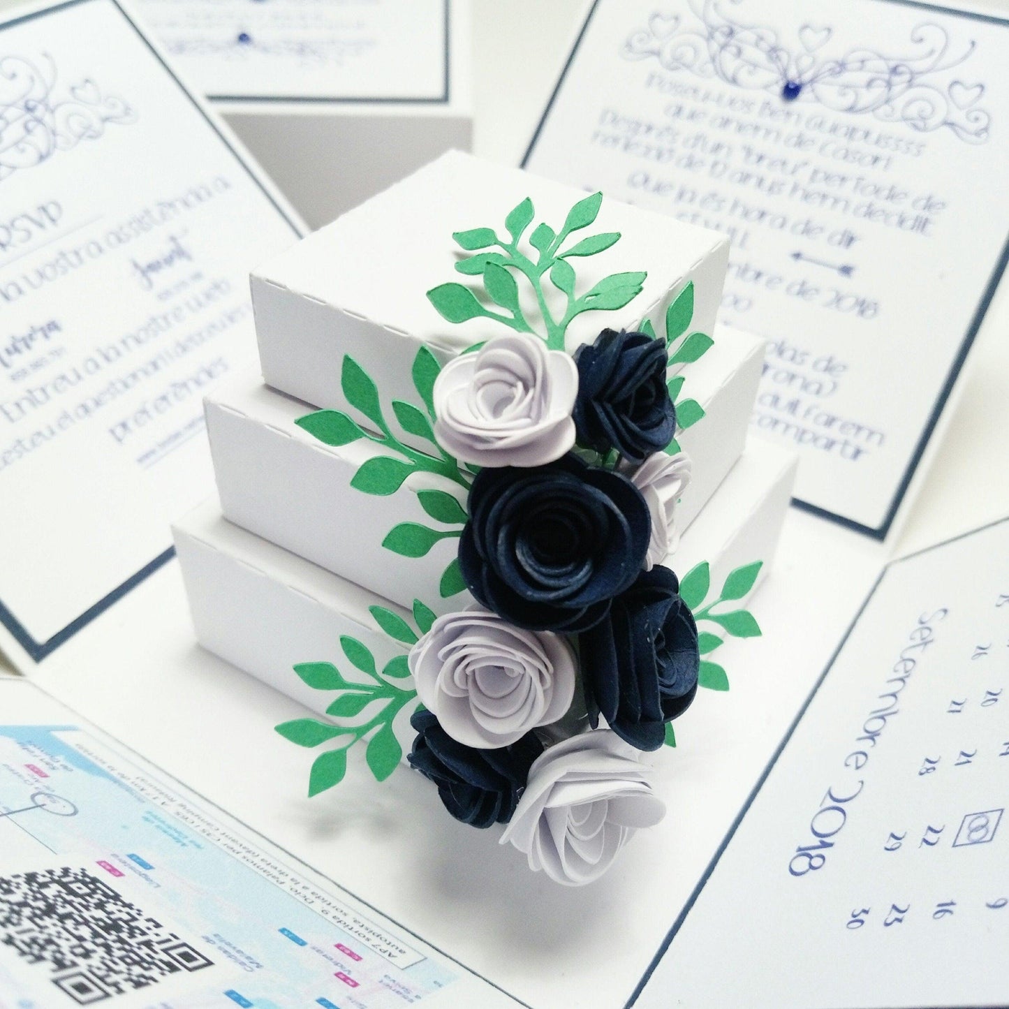 Exploding Wedding Invitation Box featuring stacked white boxes adorned with paper flowers, highlighting a three-tier decorated wedding cake centerpiece for bespoke invites and luxury wedding stationery.
