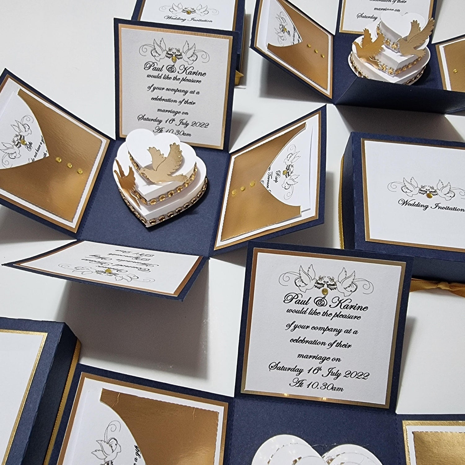 Navy Blue & Gold Exploding Wedding Invitation Boxes featuring intricate text, heart-shaped box with gold bird, and detailed cards displaying wedding details and decorative elements, including doves and gold accents.